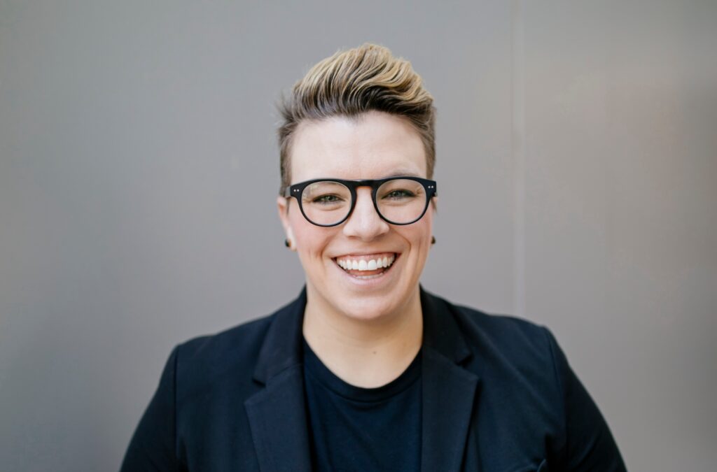 Katherine Lees smiling toward the camera. Her hair is short with a swoop and she’s wearing black-framed glasses.