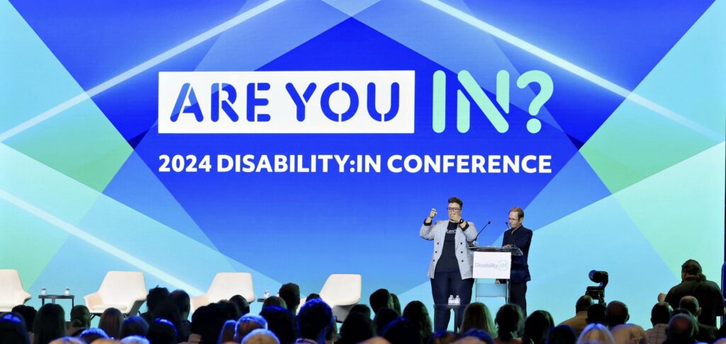 Katherine speaking at a conference on stage. Behind her, a blue screen with the words "Are You In? 2024 Disability:In Conference."